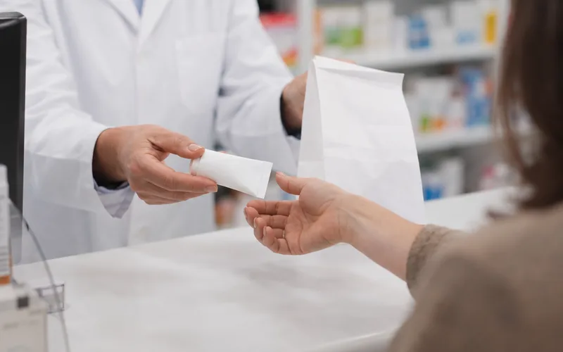 Woman's hand picking up a plain prescription cream tube and pharmacy bag at a counter