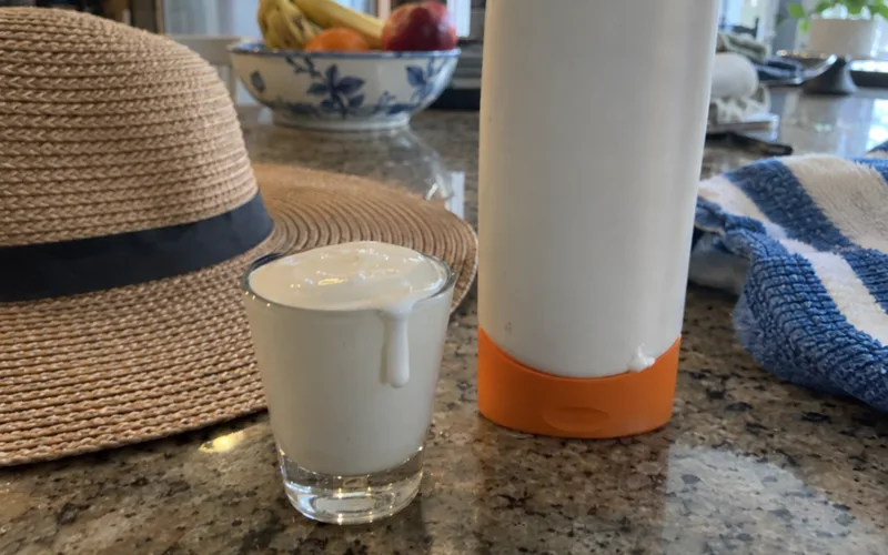 A shot glass filled with sunscreen lotion next to the bottle on a kitchen counter