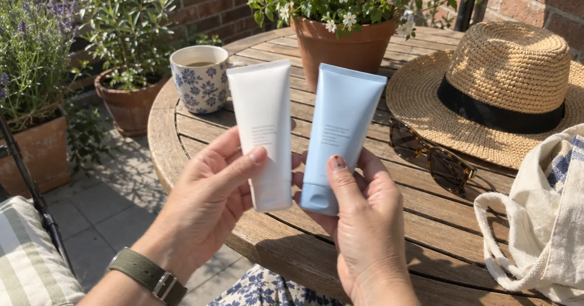 Hands comparing two sunscreen tubes on a sunny patio table beside a straw hat and sunglasses
